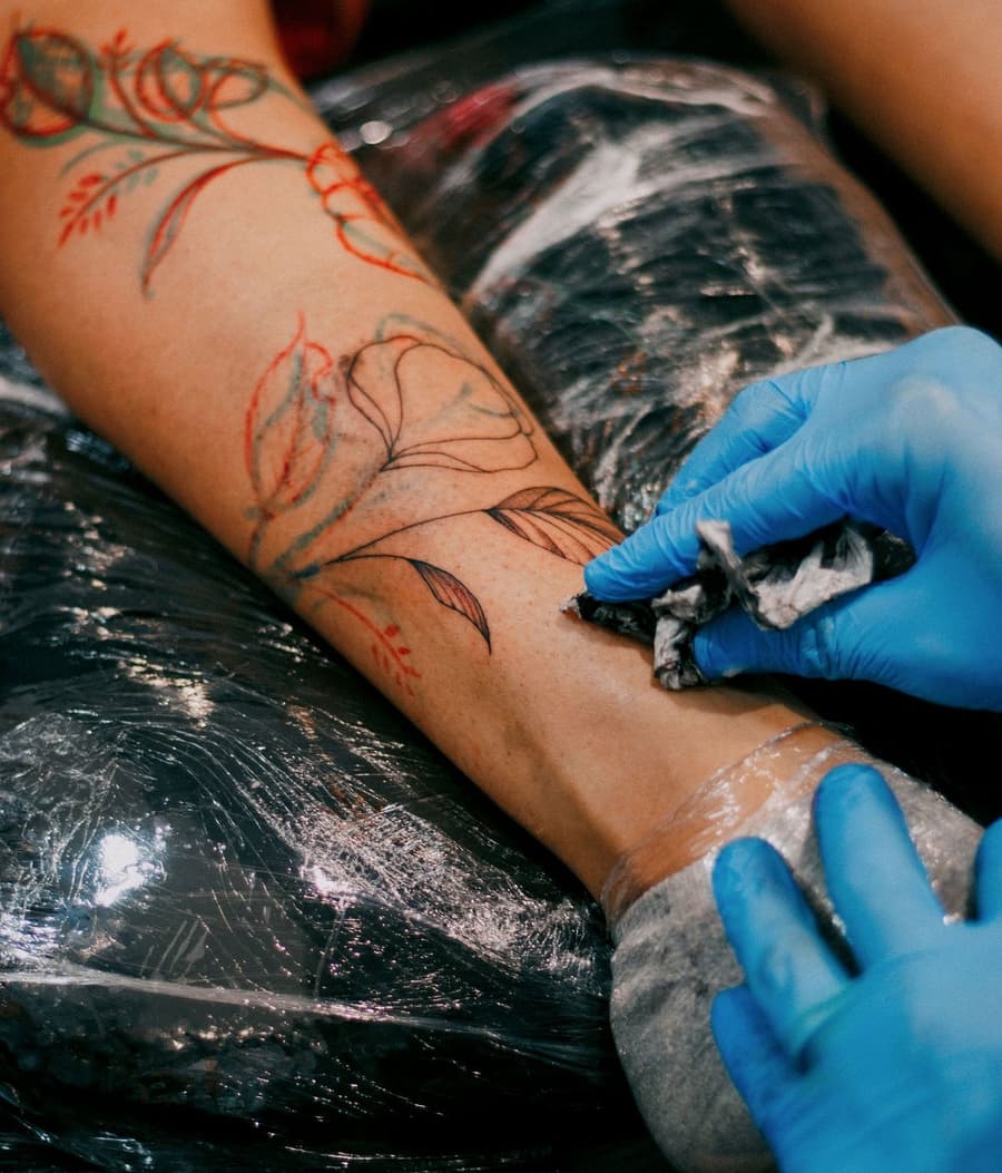 Red floral stems being tattooed along a forearm by gloved hands