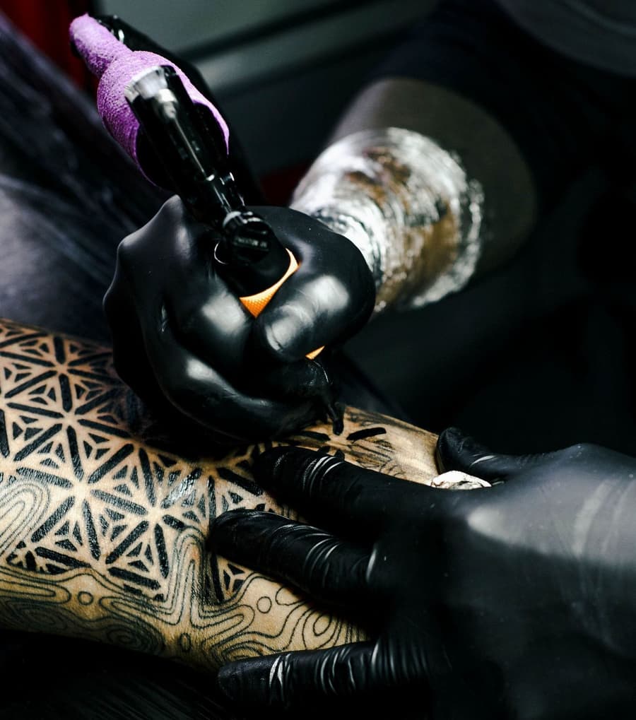 Gloved hands tattooing a flower of life pattern along a forearm