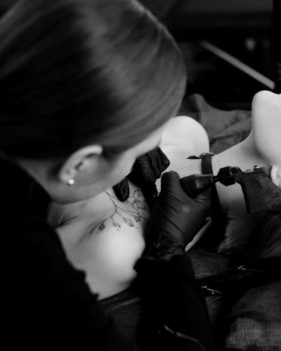 Mira Kovač tattooing a client's shoulder under a lamp, in black and white